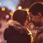 A young couple bundled in coats press foreheads together while smiling, with evening lights in the background.