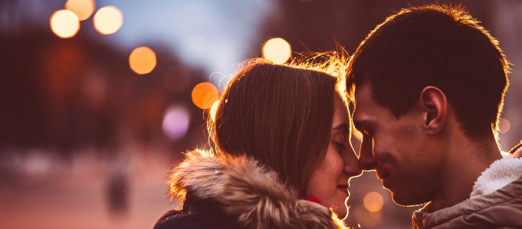 A young couple bundled in coats press foreheads together while smiling, with evening lights in the background.