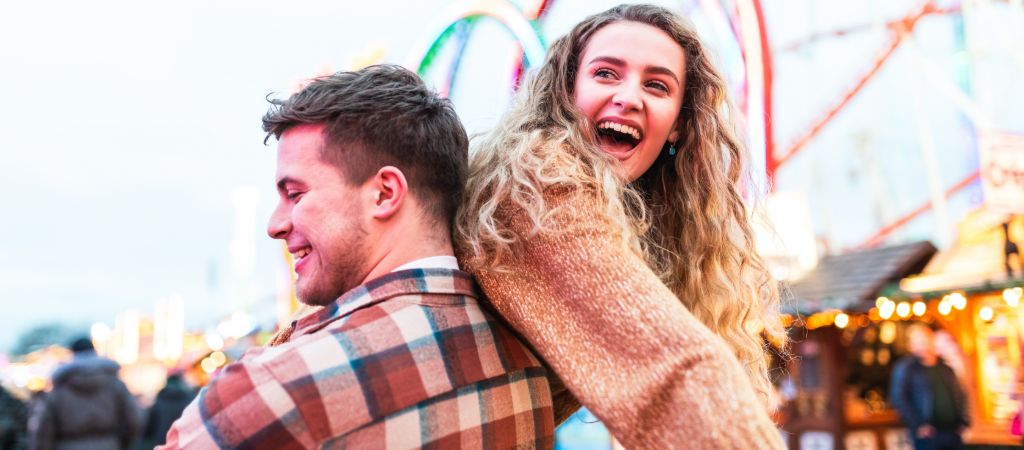 A young man carries a young woman, both smiling and laughing, at a theme park.