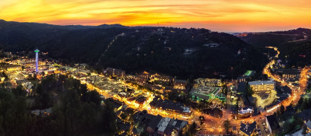 A aerial view of downtown Gatlinburg with tons of lights, the surrounding Great Smoky Mountains, and colorful oranges and pinks at sunset.