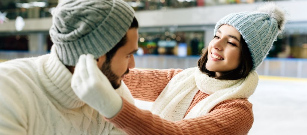A woman adjusts the toboggan on a man's head while smiling at an indoor ice-skating rink.