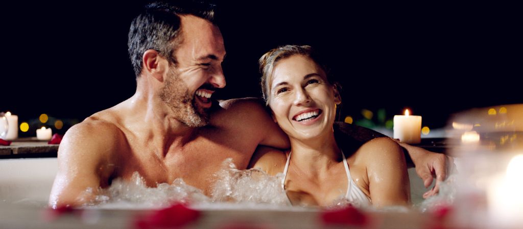 A man and woman laugh together while soaking in a jetted bathtub, with candles and rose pettles all around.