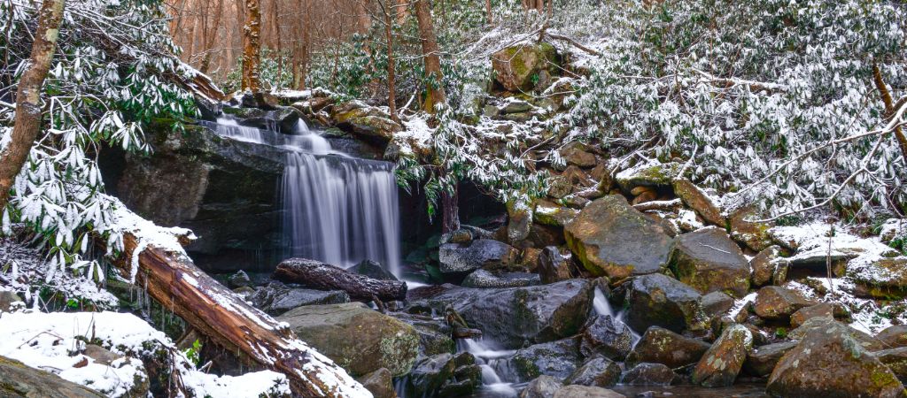 A winter scene of Rainbow Falls in the Smokies, with snow on the moss-covered stones and pine trees all around.