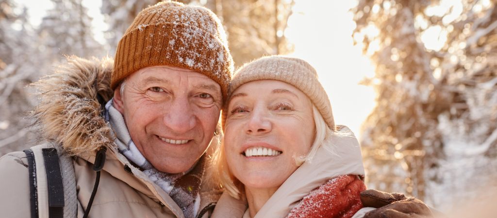And older couple bundled in winter clothing smile for a selfie together, with snow on their hats and scarves and snowy trees in the background.