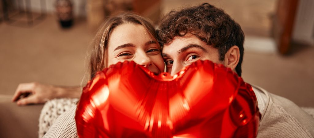 A young couple peek from behind a red hear-shaped balloon, with a smile in their eyes.