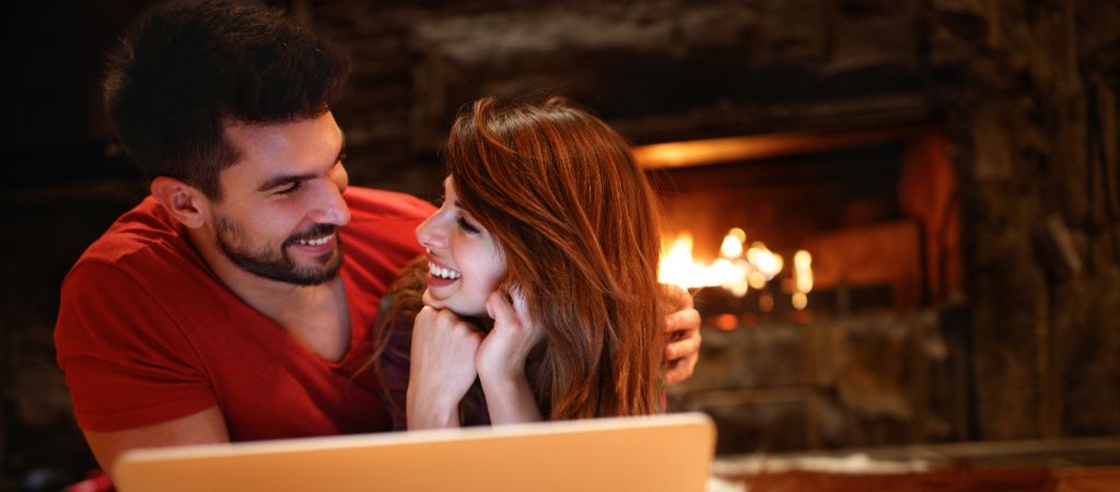 A couple laying next to each other and gazing into each other's eyes, with a laptop open in front of them and a roaring fireplace in the background.